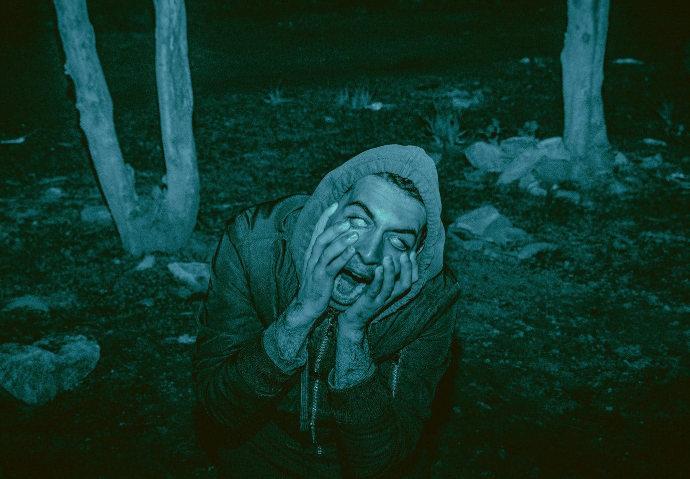 The essay's original photograph: a man in a hooded jacket kneeling in dark woods at night, hands pressed to his face